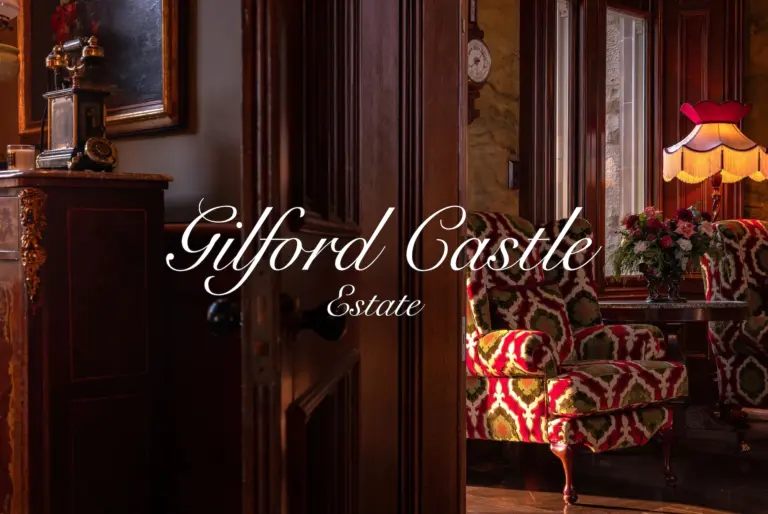 Gilford Castle Estate logo over an atmospheric interior with wood panelling, antique furniture, patterned armchairs, flowers, and warm lamplight.