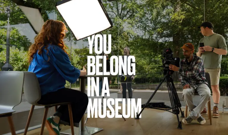 Film crew records a woman seated in Ulster Museum’s café beneath the words “You Belong in a Museum”.